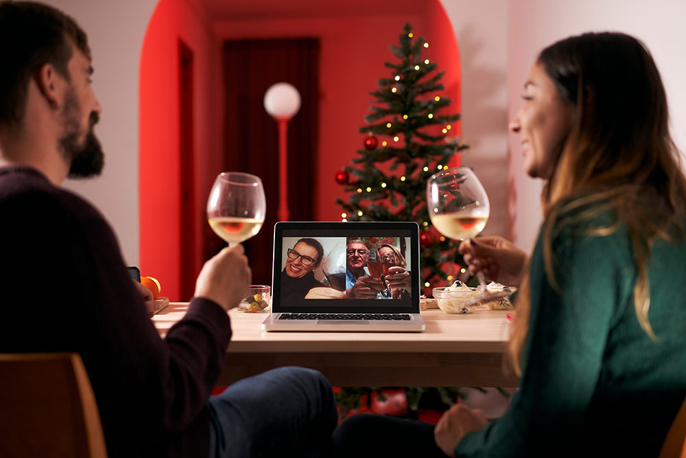 Young couple in front of Christmas tree on video call with senior family members having dinner and drinks celebrating the holidays